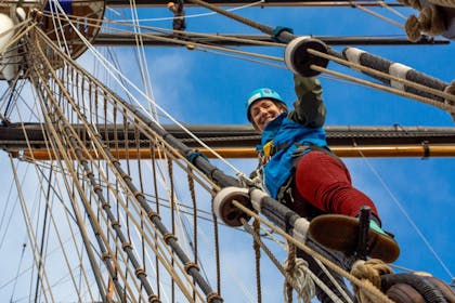 A smiling woman in protective clothing climbing the Cutty Sark rigging under a bright blue sky